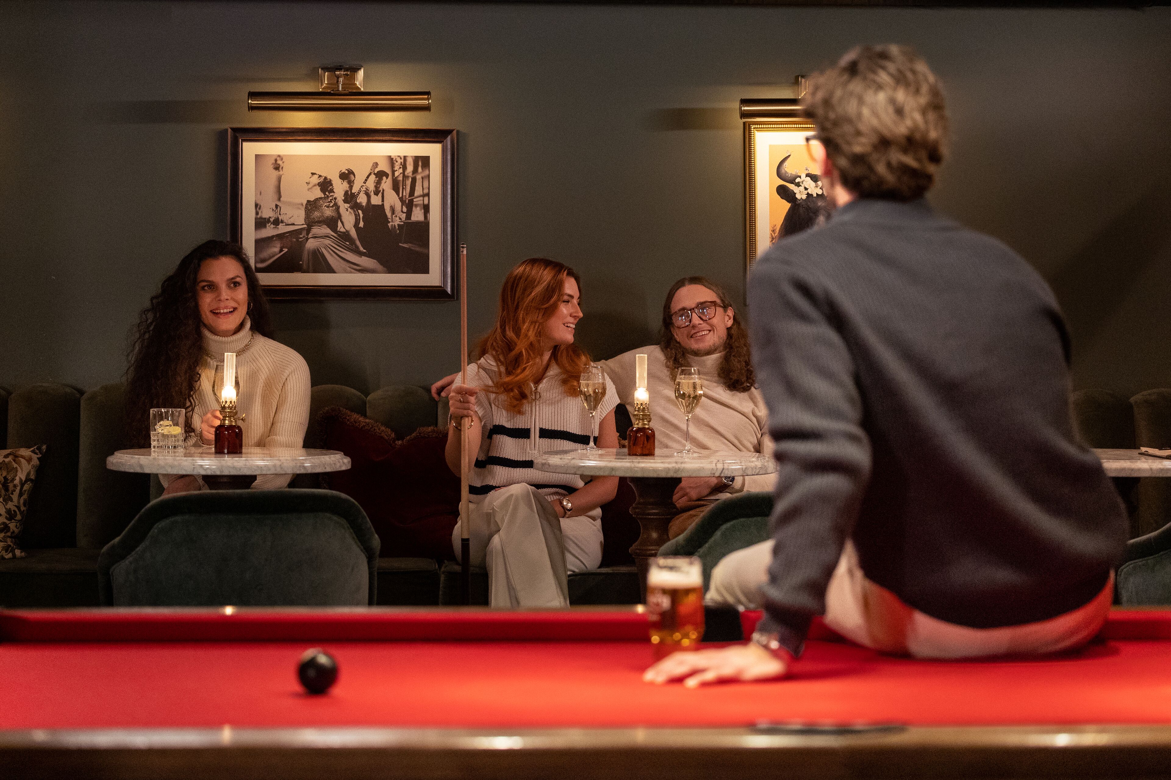 A billiards table with four people seated around it, a framed picture on the wall, and a pool table in the foreground.