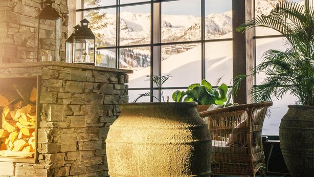 A stone fireplace with a log fire, large potted plants, wicker furniture, and a view of mountains through large windows. - Fyri Resort Hemsedal (Hemsedal)