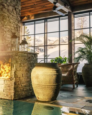 Una chimenea de piedra con fuego de leña, grandes macetas con plantas, muebles de mimbre y vistas a montañas a través de amplias ventanas.