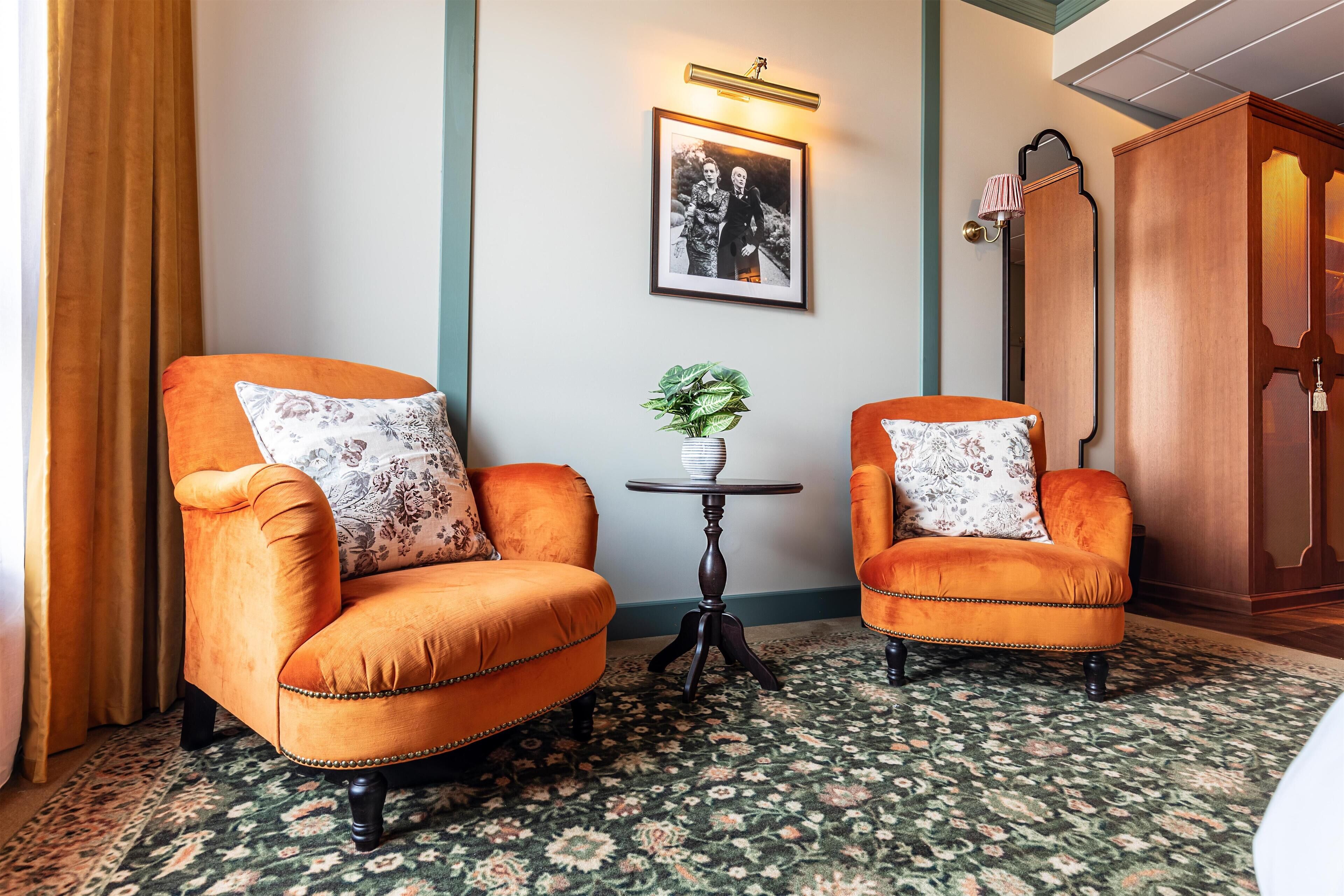 A room with two orange armchairs, a small table with a plant, a framed photograph on the wall, and a wooden cabinet.