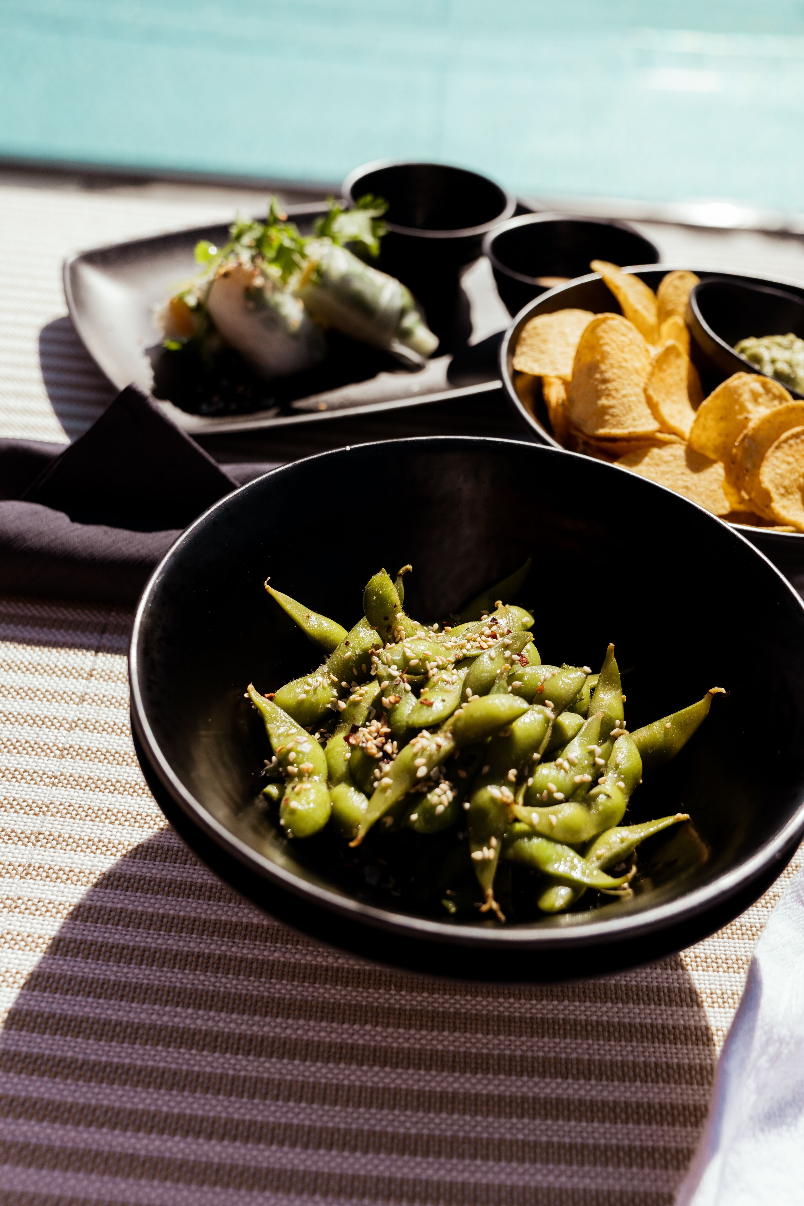 A table with a bowl of edamame, a plate of spring rolls, and a bowl of chips.