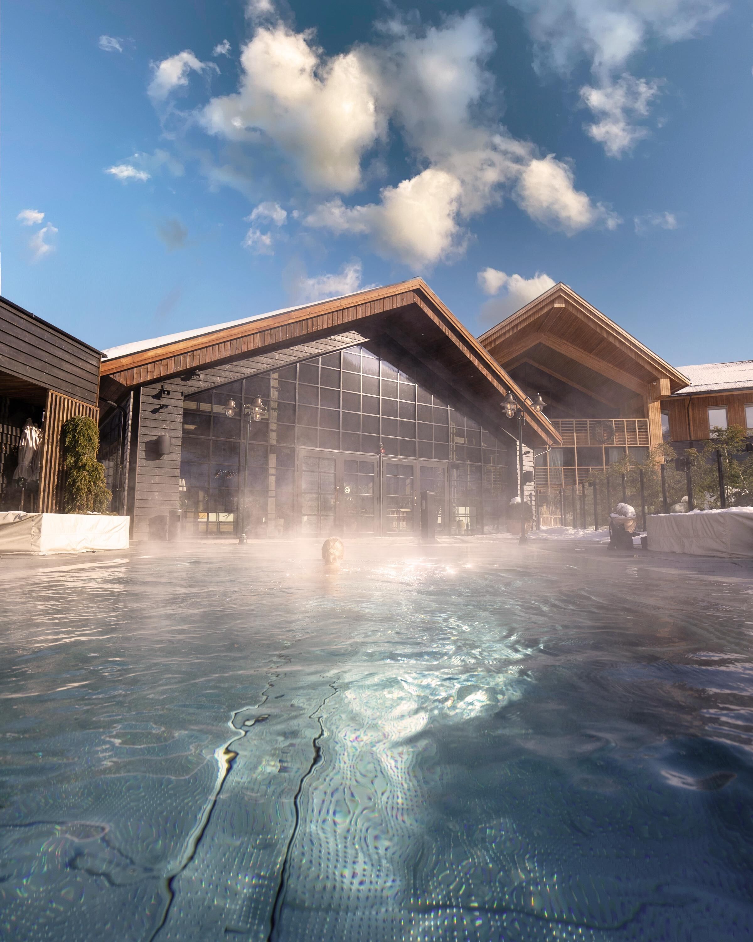 Un espace de bien-être comprenant un grand bâtiment à structure de verre, une piscine d’eau thermale et une construction en bois dotée d’un toit en pente.