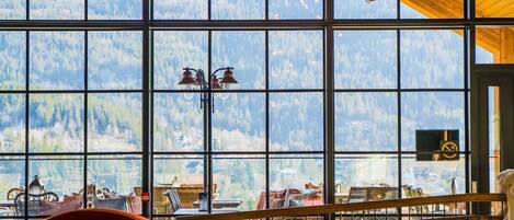 A dining area with a long table set for a meal, large windows offering a view of a mountainous landscape, and a wooden ceiling.