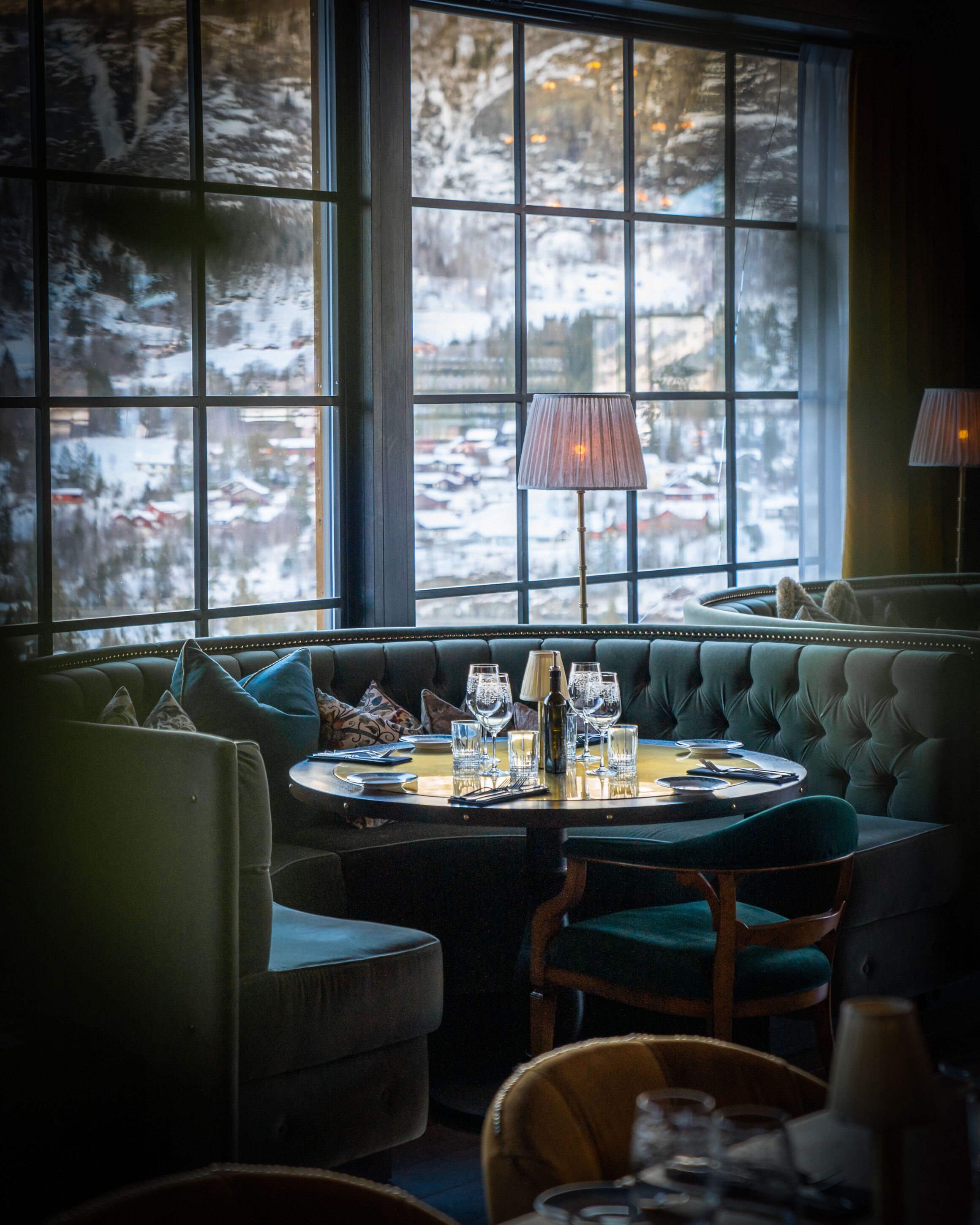 Un rincón acogedor con una mesa redonda dispuesta para comer, ventanas grandes con vista nevada y una lámpara sobre la mesa.
