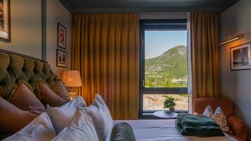 Quarto de hotel com cama, poltrona, abajur, janela com cortinas e vista para uma montanha.