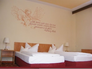 Quarto de hotel com duas camas, cabeceira vermelha e decoração de parede com um anjo alado.