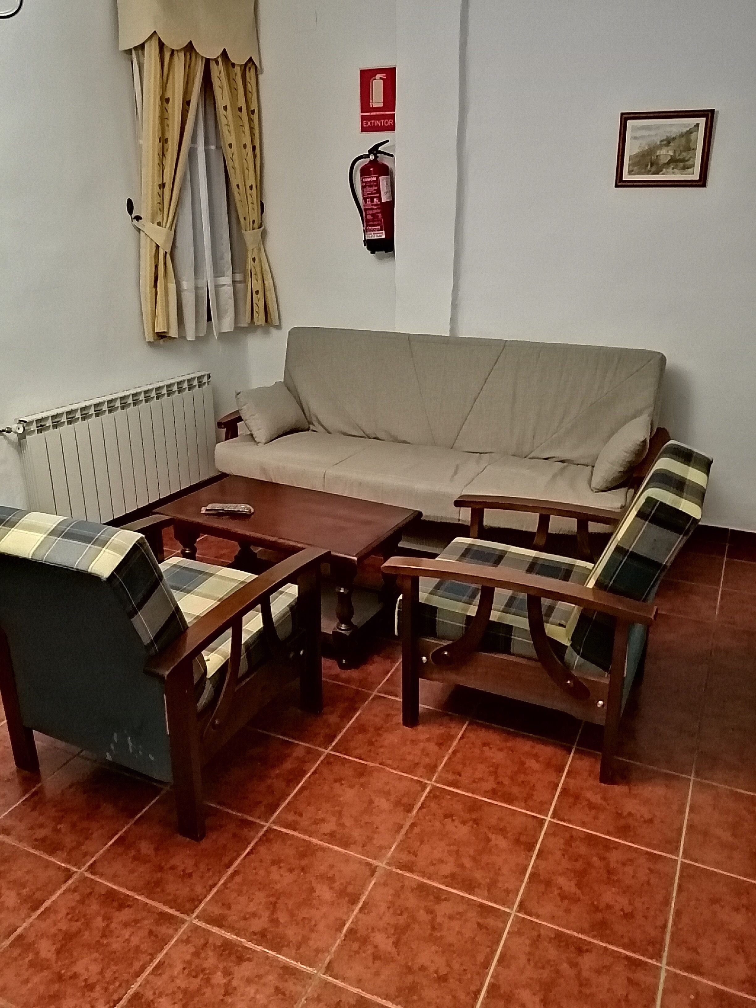 Un salon comprenant un canapé, deux fauteuils, une table basse, un radiateur, des rideaux, un extincteur et un tableau encadré accroché au mur.