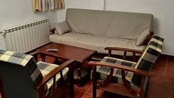 Un salon comprenant un canapé, deux fauteuils, une table basse, un radiateur, des rideaux, un extincteur et un tableau encadré accroché au mur.