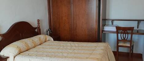 A bedroom with a wooden wardrobe, a bed with a white bedspread, a wooden chair, and a small table.