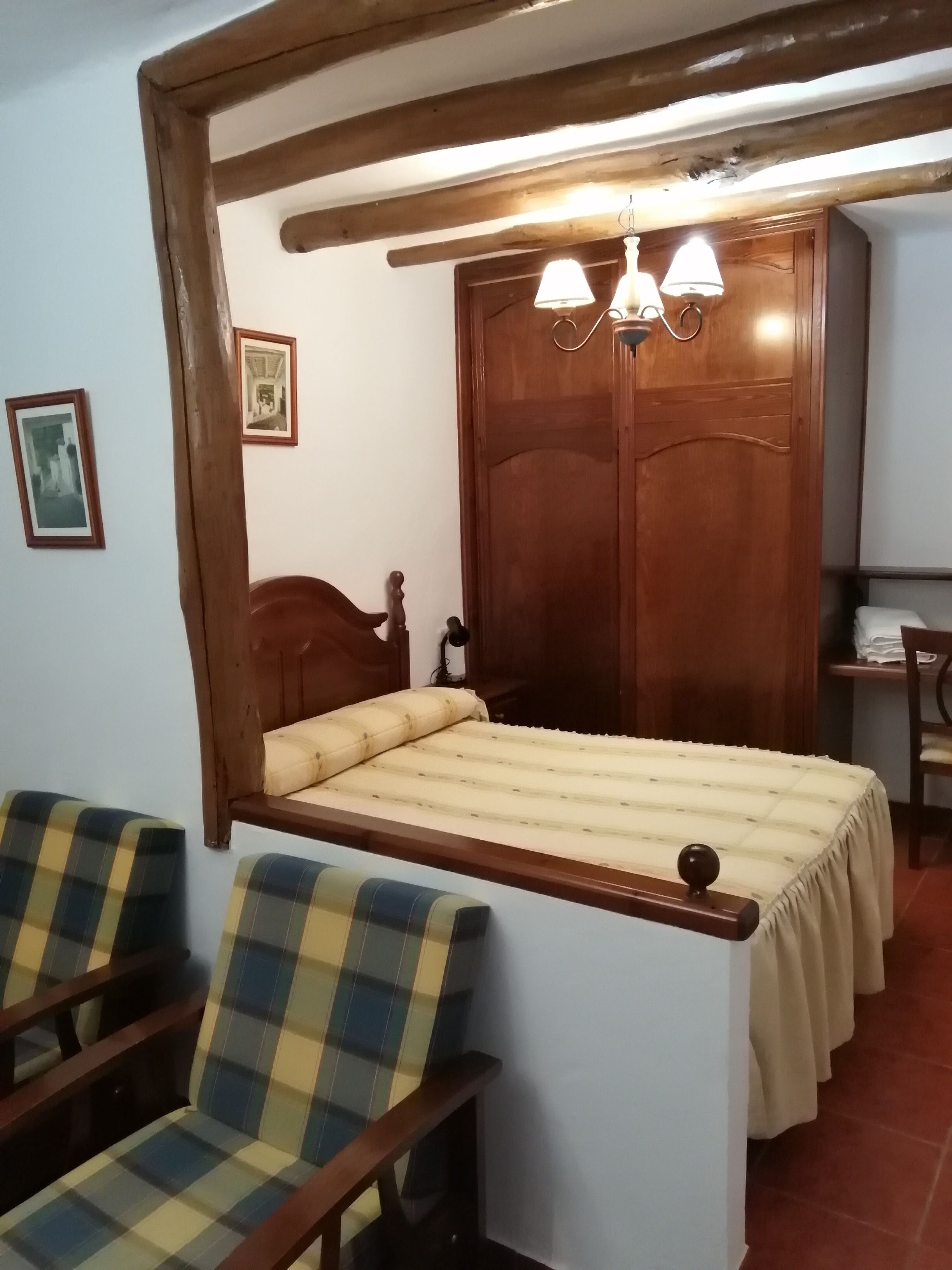 A bedroom with a wooden bed, two plaid chairs, a wooden wardrobe, and a desk with a lamp.