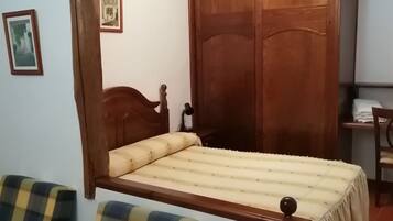 A bedroom with a wooden bed, two plaid chairs, a wooden wardrobe, and a desk with a lamp.