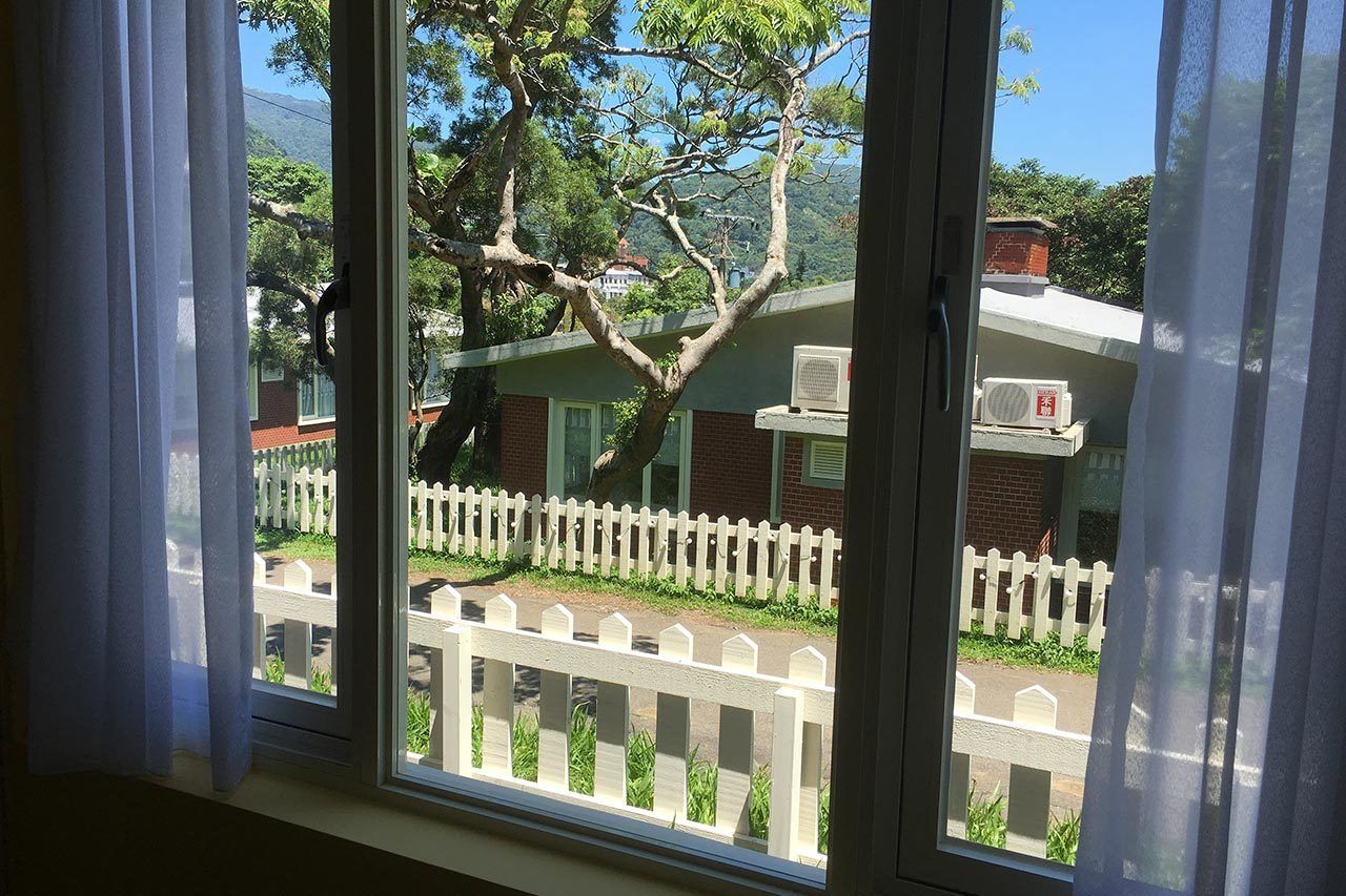 A window with a view of a white picket fence, a tree, and a building with a brick facade.