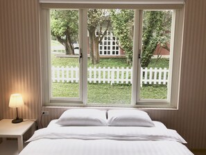 A bedroom with a bed, a nightstand, and a window with a view of a white fence and trees. - American Village Resort (Taipei)