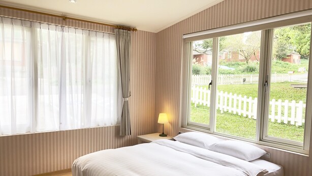Et soveværelse med et stort vindue, et natbord med en lampe, og en seng med hvide sengetøj.