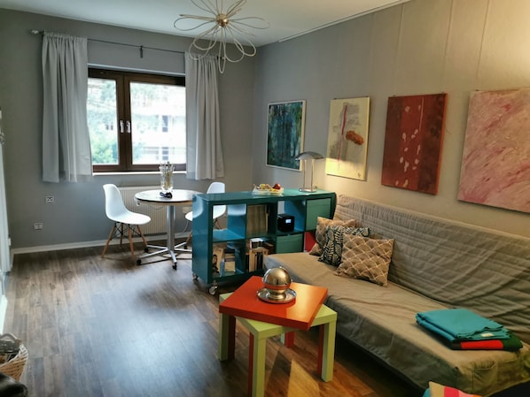 A modern living room with a sofa, a coffee table, and a dining table. There are paintings on the wall and a window with curtains.