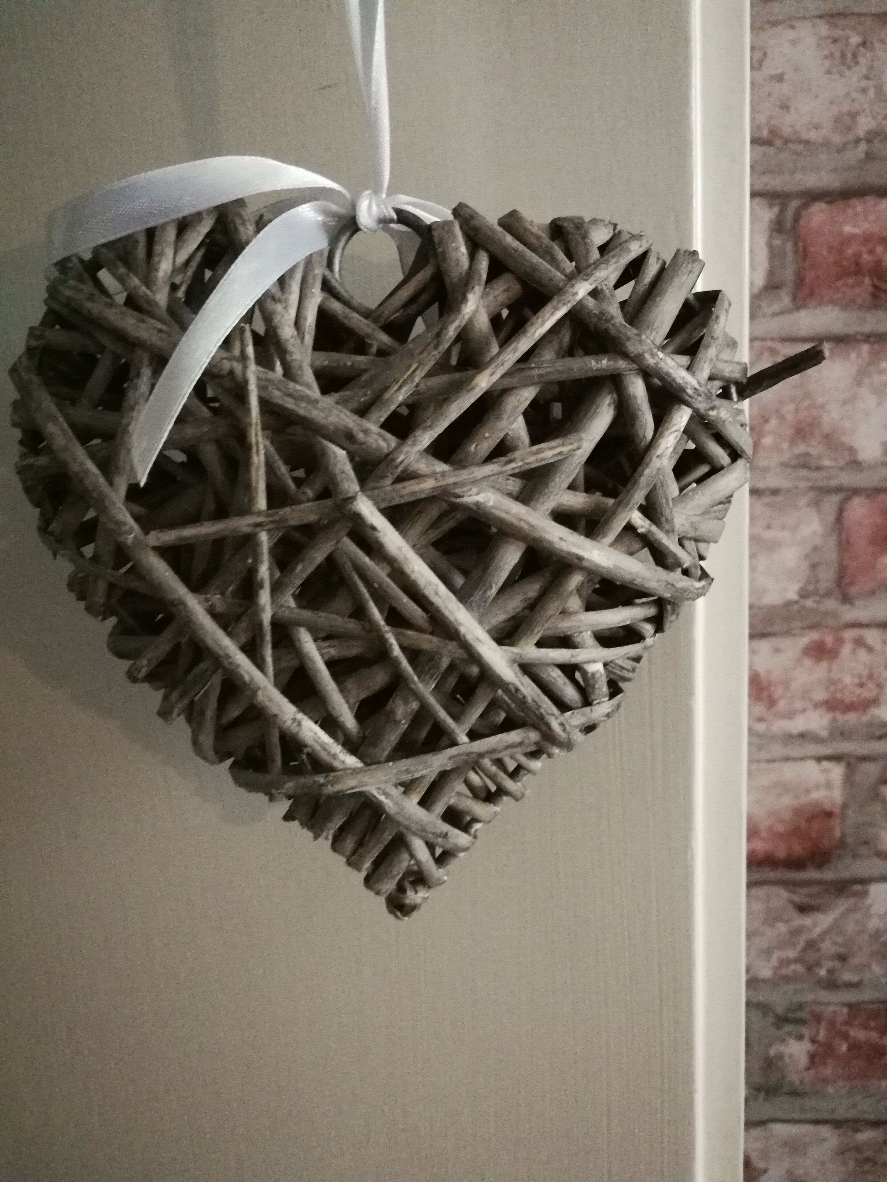Ein herzförmiges Dekorationsobjekt aus verschlungenen Ästen, gebunden mit einem weißen Band, hängt an einer Wand mit Ziegelmuster.