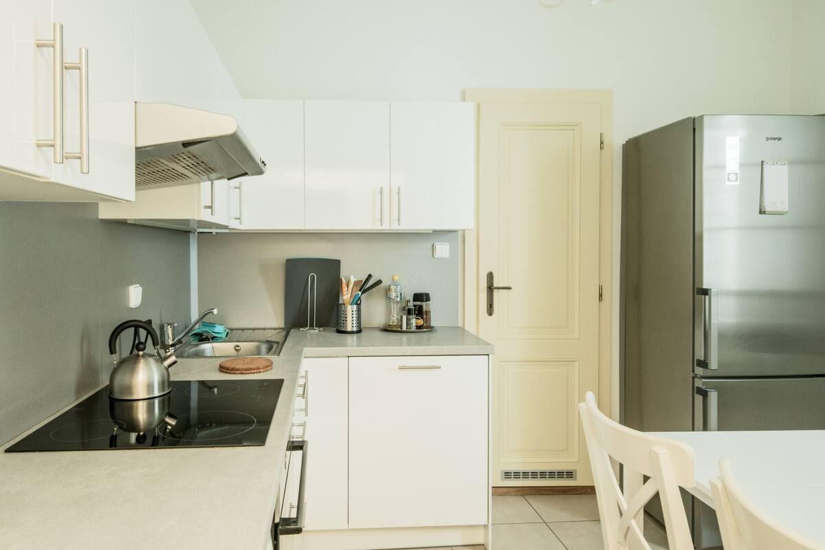 A compact kitchen with white cabinets, a stainless steel refrigerator, a kettle, and a small sink.