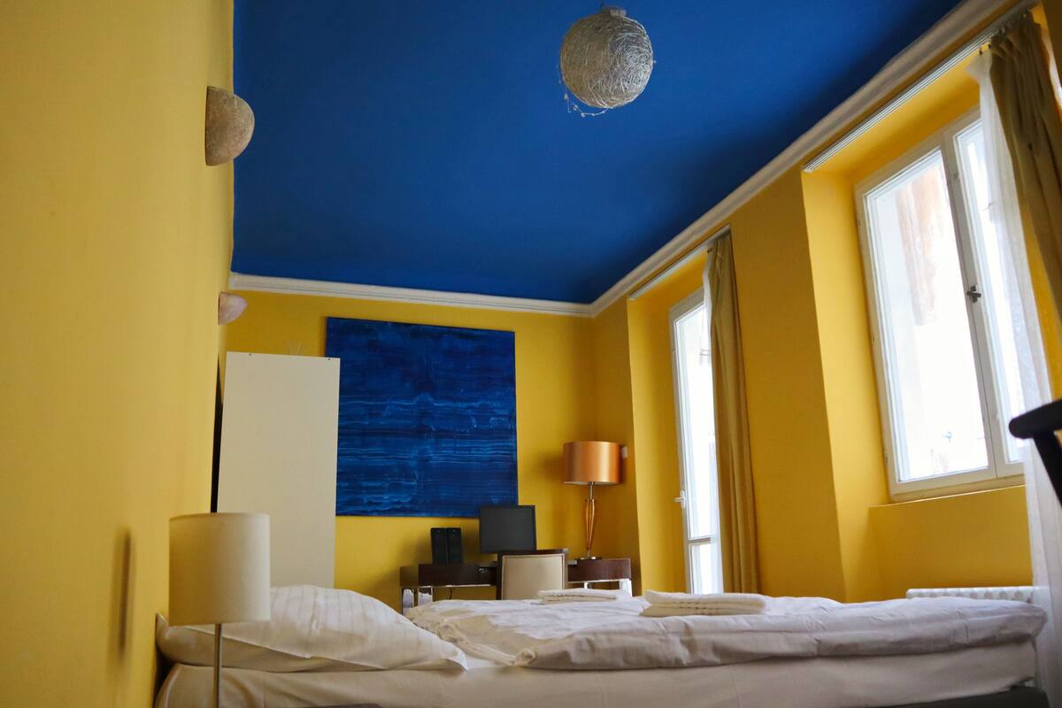 A hotel room with a bed, bedside table, lamp, and a large blue artwork on the wall.