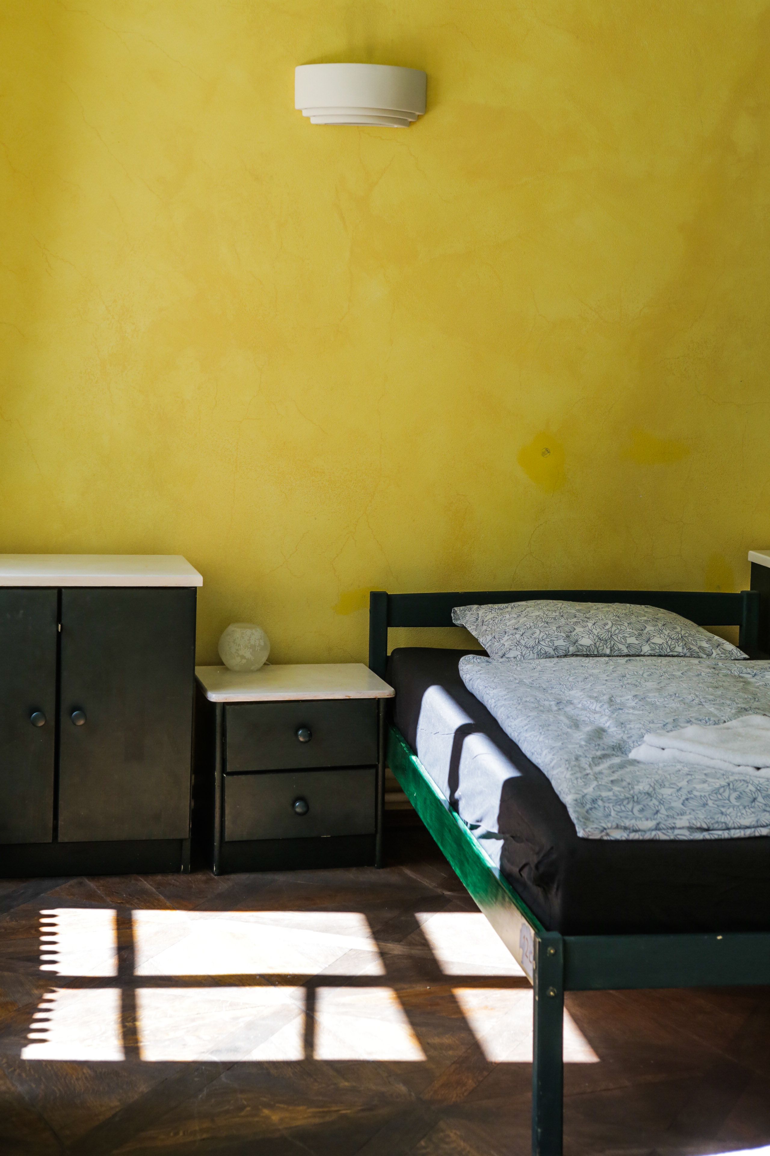 Quarto com cama, mesa de cabeceira, cômoda e luminária fixa na parede.