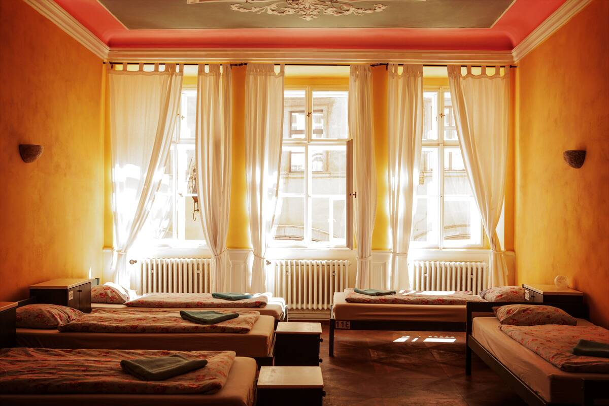 A hotel room with four beds arranged in a row, a radiator, and a window with curtains.