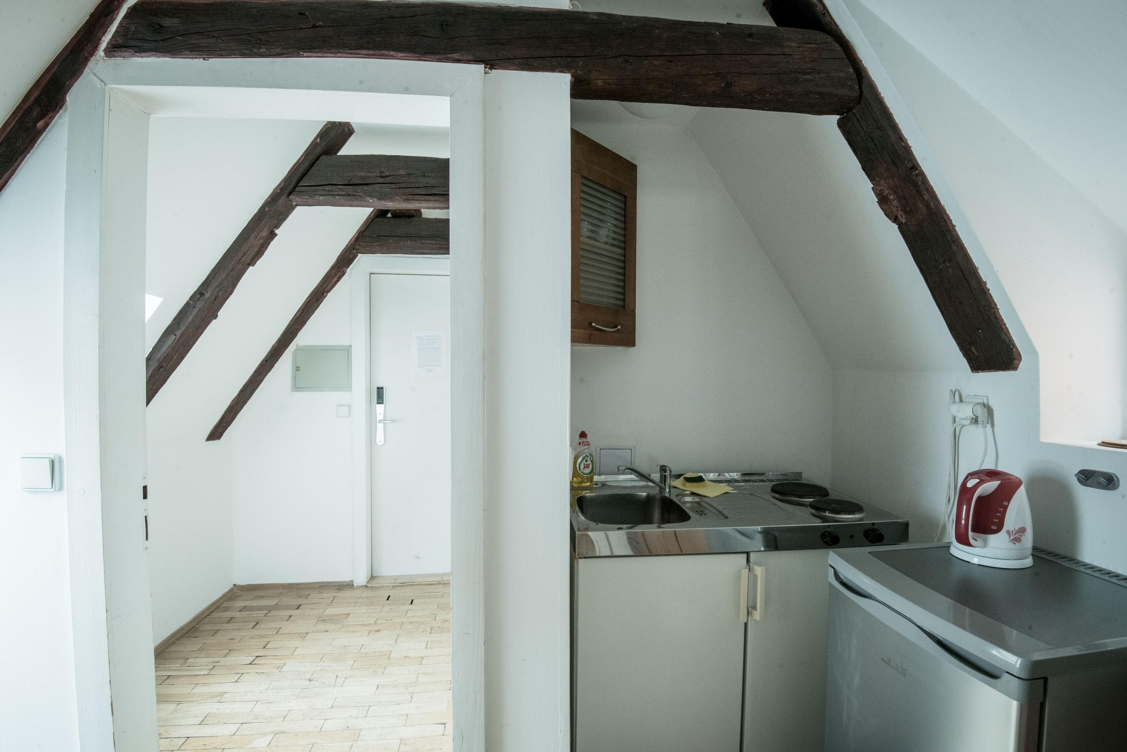 A kitchen with a stove, sink, and kettle, wooden beams on the ceiling, and a door leading to another room.