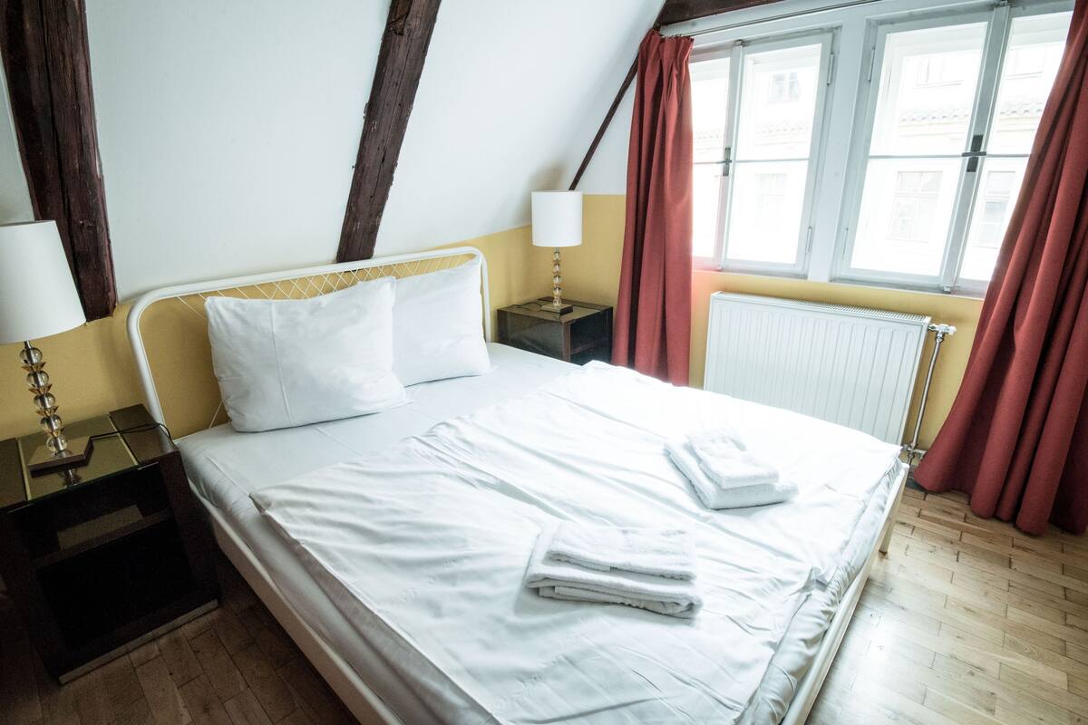 A neatly made bed with white linens and towels, a bedside lamp, and a window with curtains.
