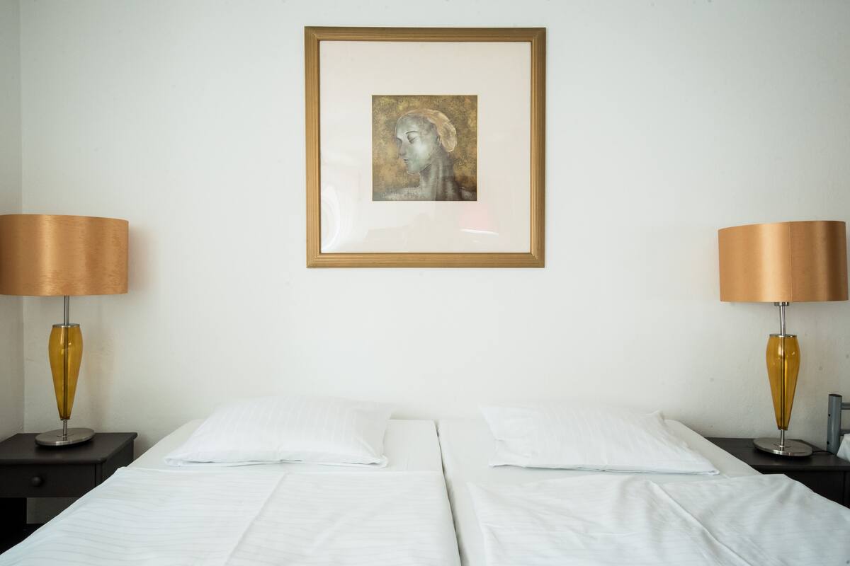 A hotel room with two beds, white linens, a framed artwork on the wall, and two lamps with golden shades.