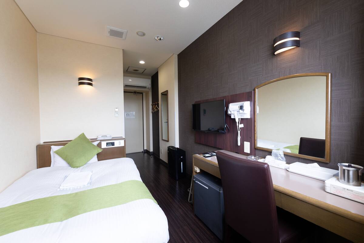 Quarto de hotel com cama, escrivaninha, cadeira, espelho e televisão.