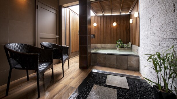 Un baño moderno con suelo de madera, dos sillas de mimbre, una zona con piedras negras y una ducha con cerramiento de vidrio.