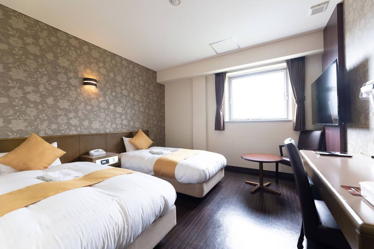 Quarto de hotel com duas camas, uma escrivaninha, uma cadeira, uma televisão e uma janela.