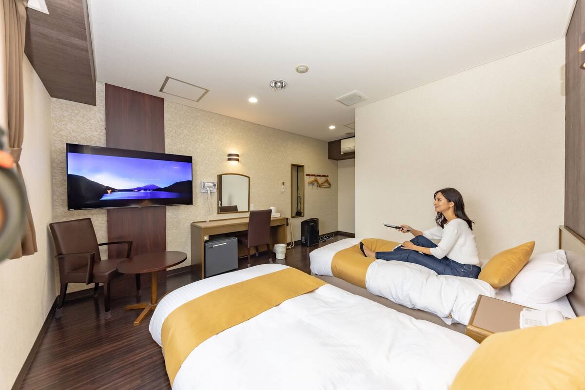 Quarto de hotel com duas camas, uma mesa de trabalho com computador, televisão e uma cadeira.