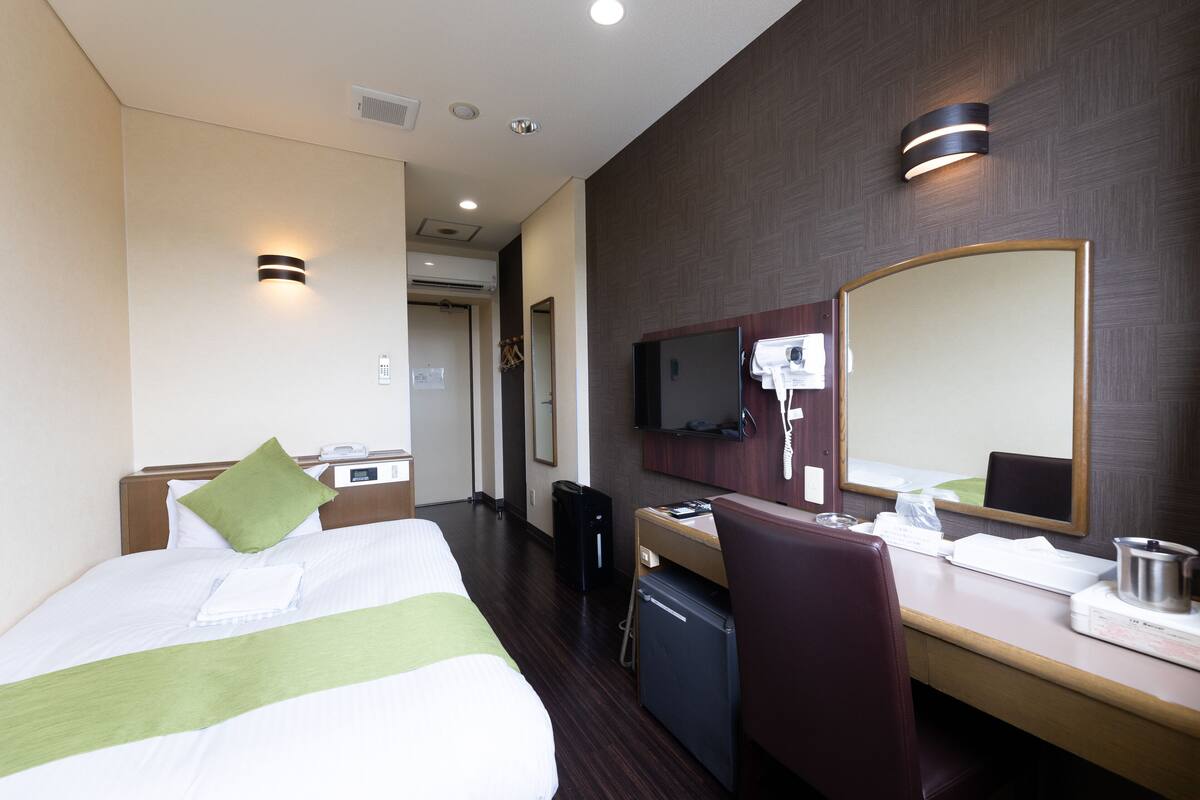 Quarto de hotel com cama, escrivaninha, cadeira, espelho e televisão.