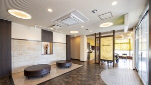 Un lobby moderno con una zona de recepción, un área de descanso con dos mesas redondas y un restaurante visible a través de una pared de vidrio.