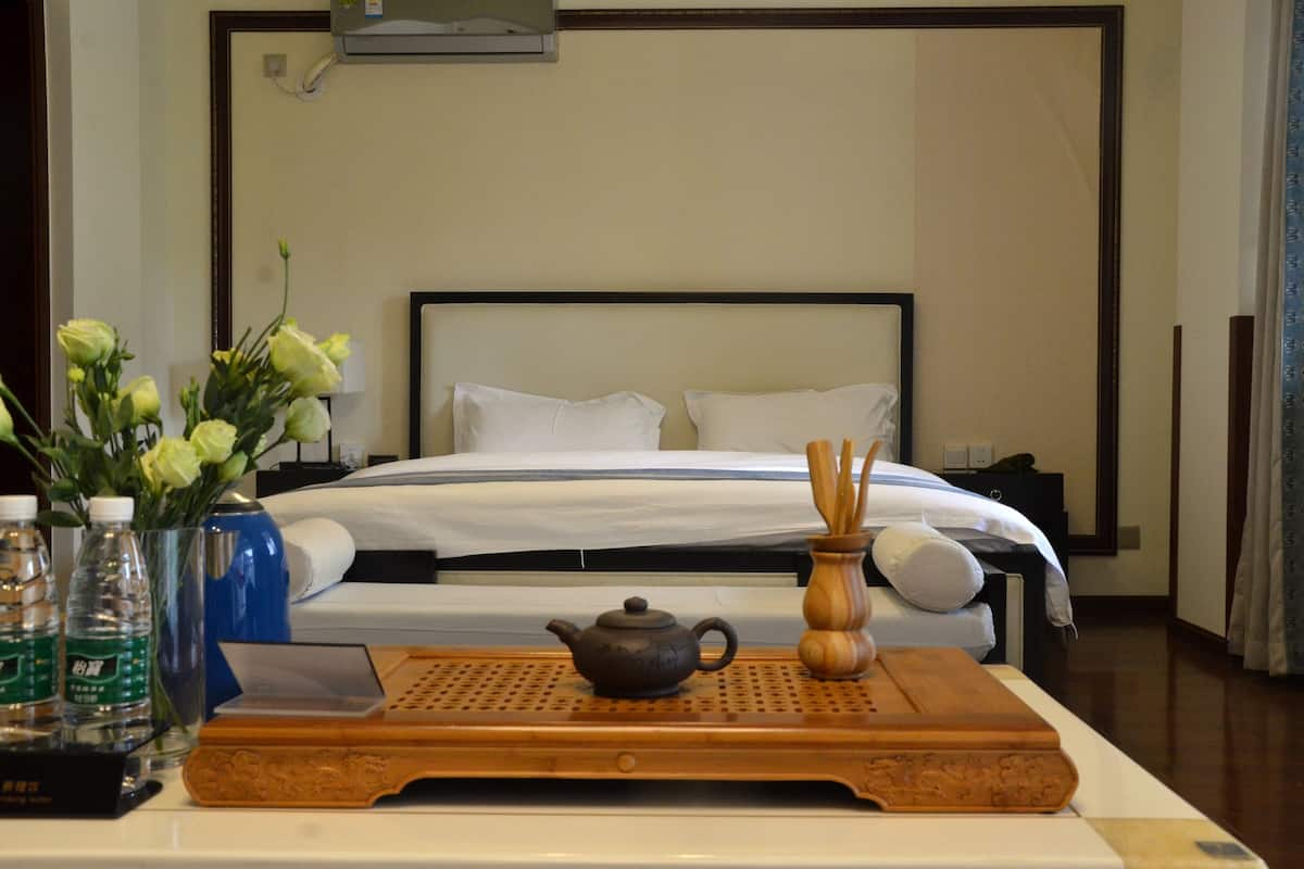 Uma cama bem arrumada com lençóis brancos, uma mesa de cabeceira com uma chaleira, um vaso com flores e uma bandeja com garrafas de água e um telefone.