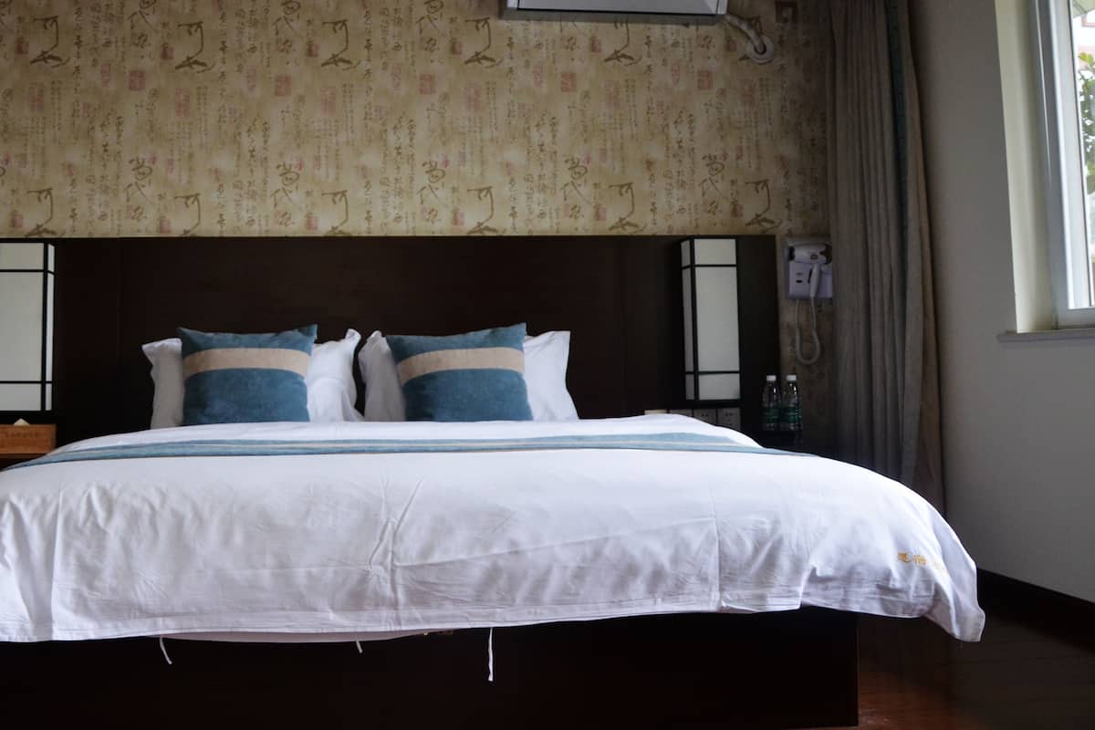 Cama bem arrumada com roupa de cama branca e azul, cabeceira escura e parede com padrões de escrita tradicional.