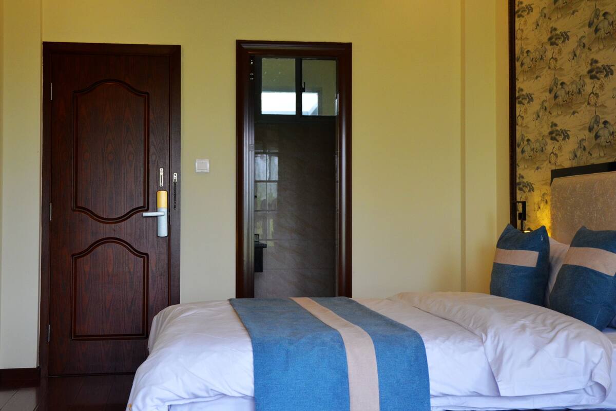 Quarto de hotel com porta de madeira, cama com colcha azul e bege, e papel de parede floral na parede.