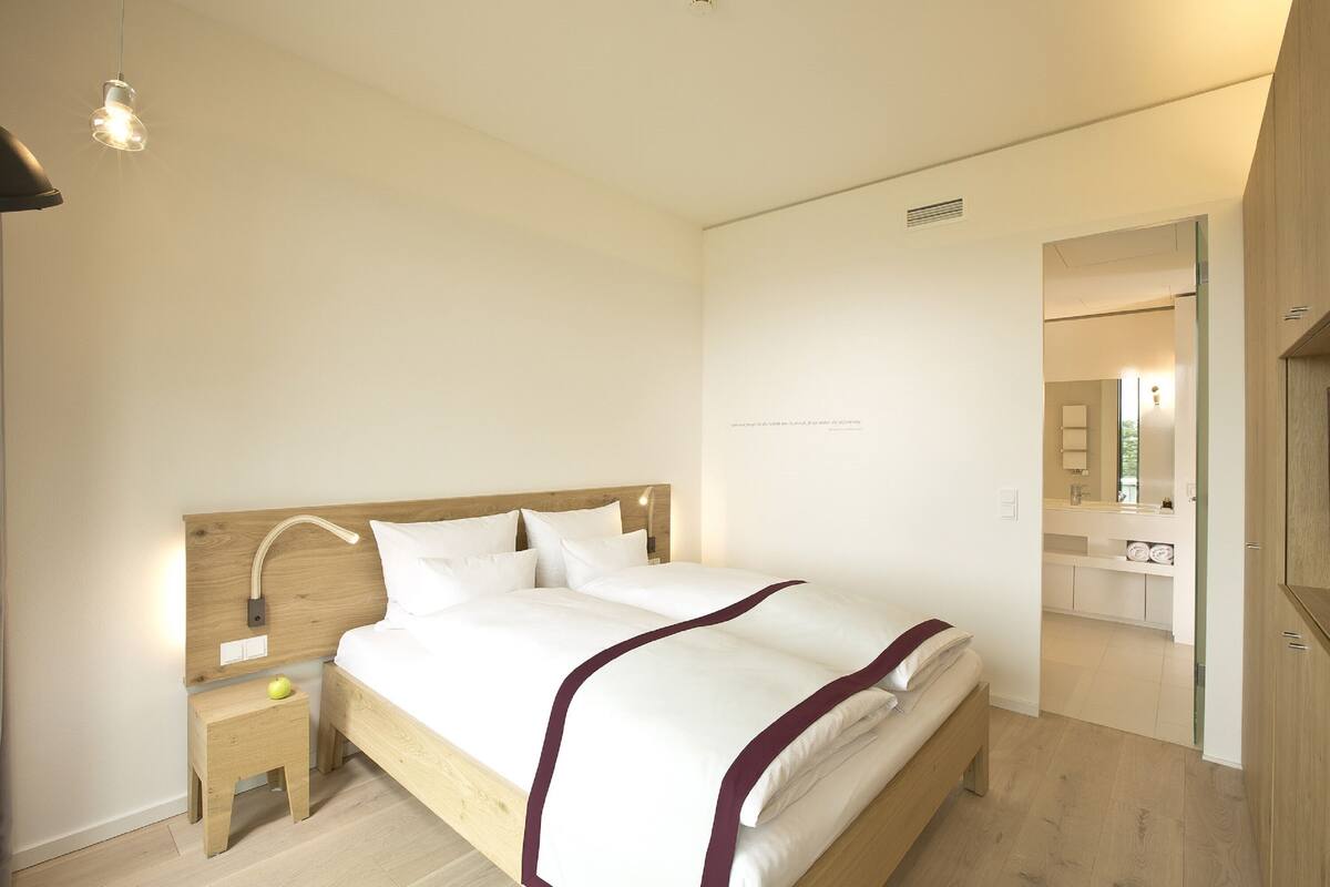 A bedroom with a wooden headboard, a bedside table with a lamp, and a view into the bathroom.