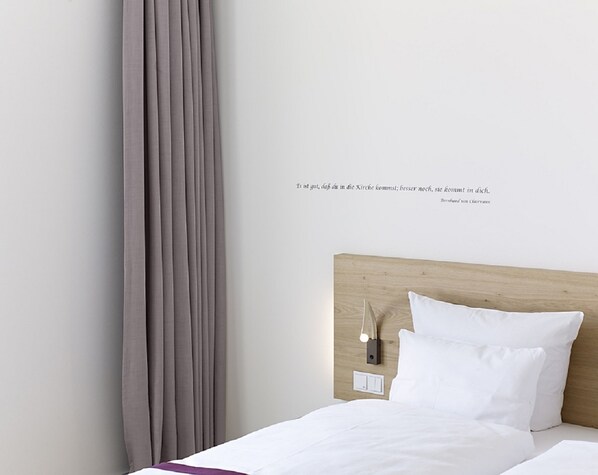 Una cama bien hecha con sábanas blancas y un cabecero de madera. Hay una lámpara de pared e interruptor sobre la cama.