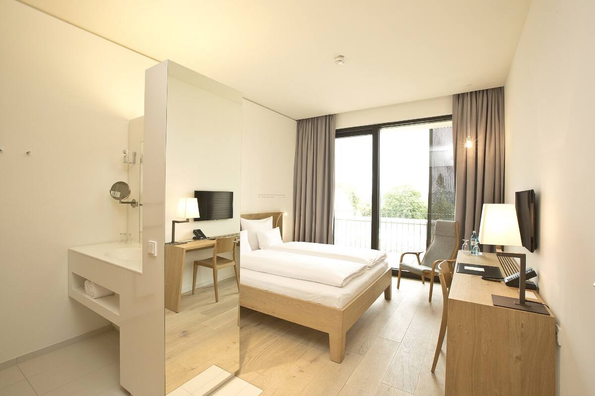 A hotel room with a bed, desk, chair, and a large window with curtains.