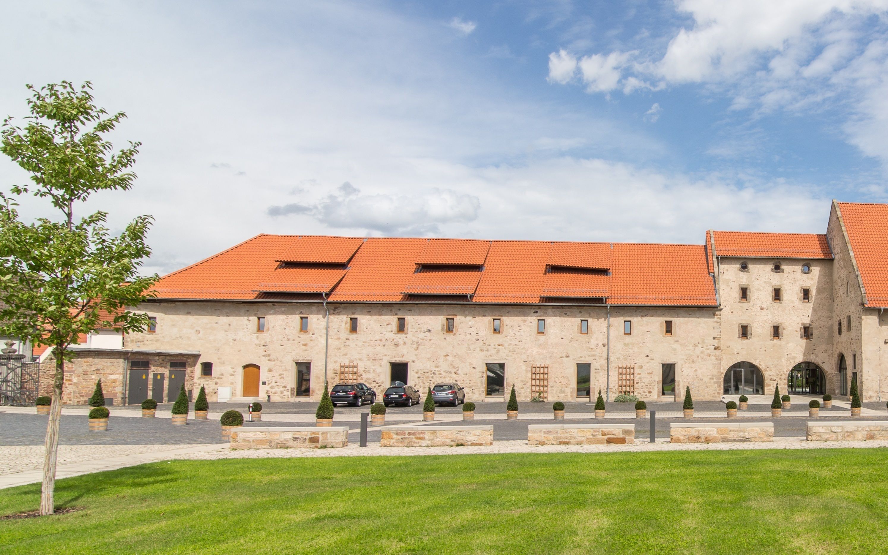 A historic building with a red-tiled roof, stone walls, and a courtyard with parked cars and potted plants.