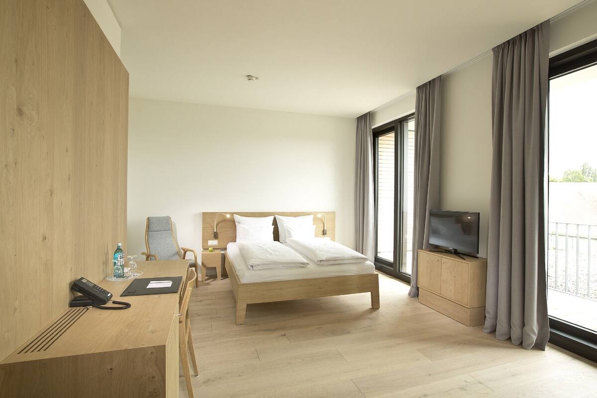 A hotel room with a wooden bed, a desk with a phone, a TV, and a large window with curtains.
