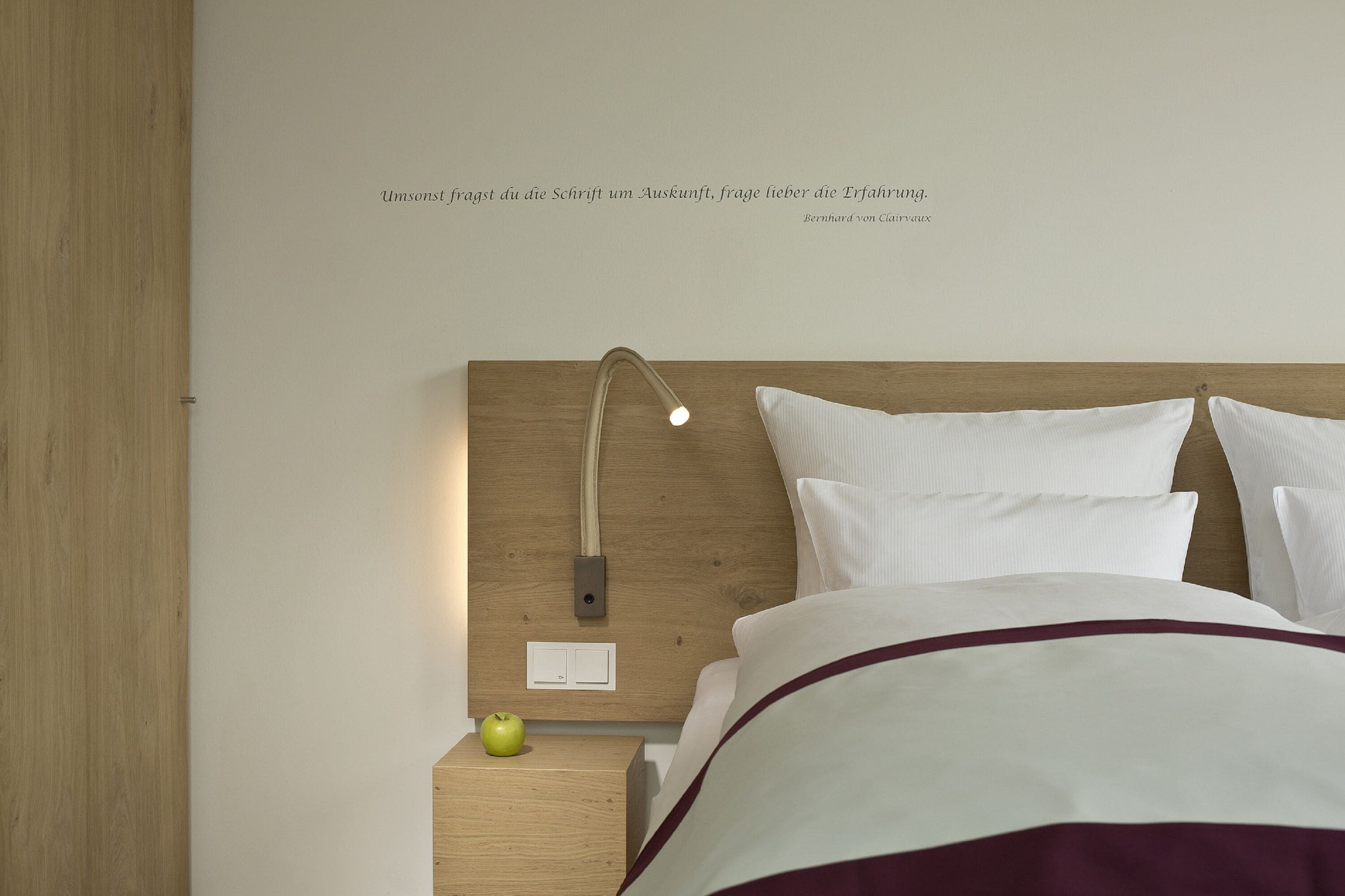 Una cama bien hecha con sábanas blancas y un detalle color burdeos, una lámpara de noche, una mesita de noche de madera con una manzana verde, y una pared con una cita en alemán.