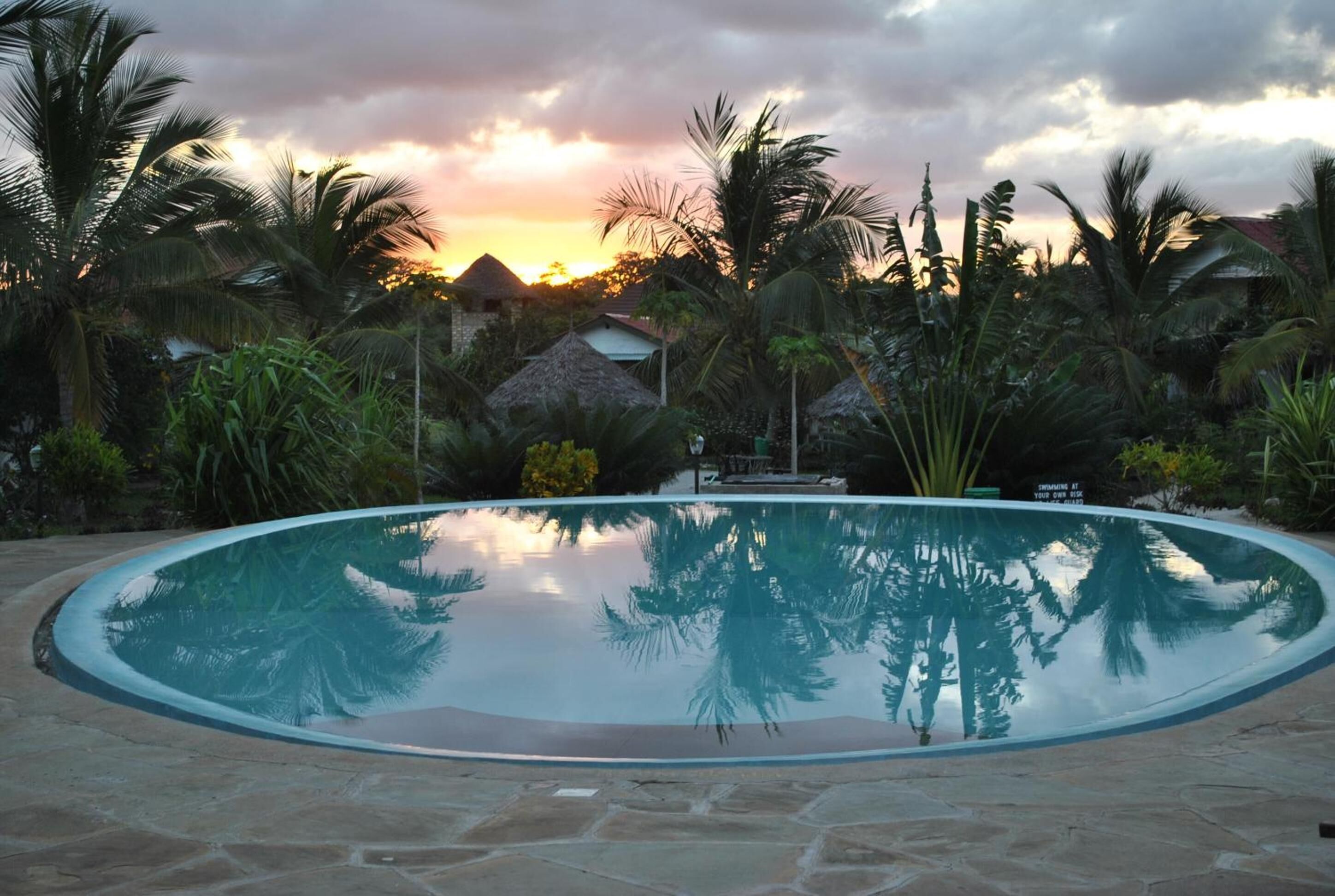 A clear swimming pool surrounded by palm trees and thatched-roof huts, reflecting the sunset.