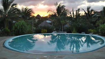 A clear swimming pool surrounded by palm trees and thatched-roof huts, reflecting the sunset.