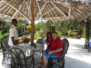 A covered outdoor seating area with a thatched roof, wicker chairs, and a table with drinks. - African Dream Cottages (Diani Beach)