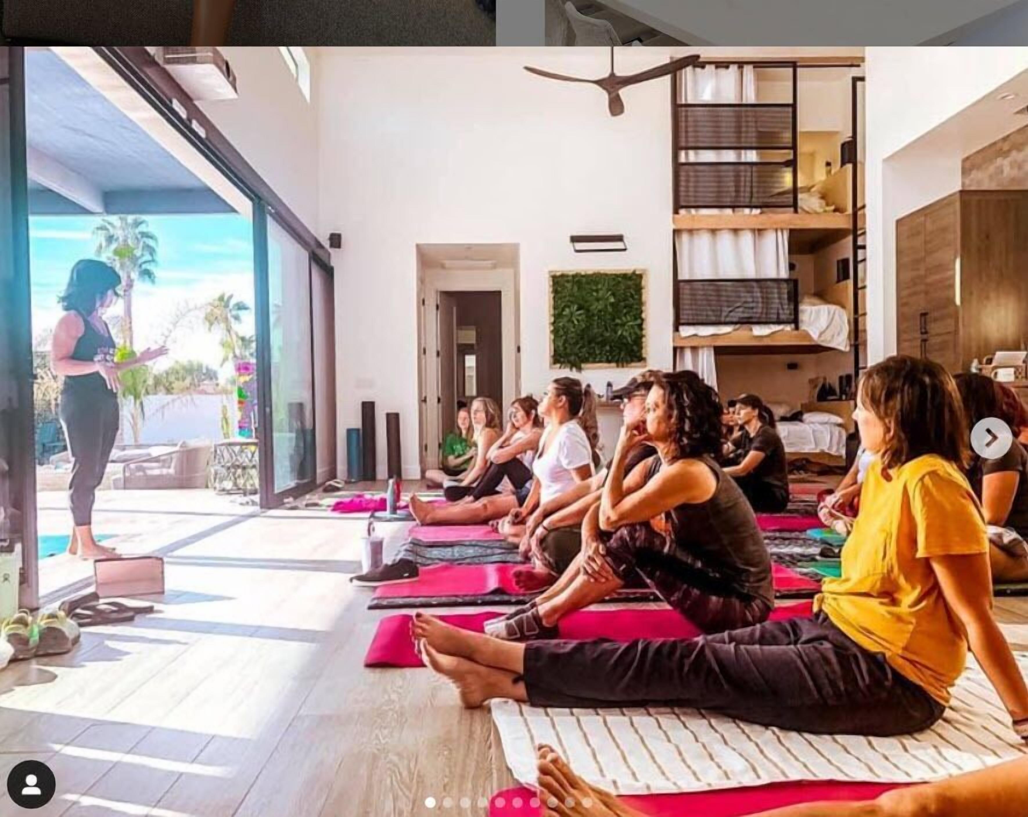Un groupe de personnes participe à un cours de yoga, assises sur des tapis dans une pièce dotée d’une grande fenêtre, d’un ventilateur de plafond et d’une plante.