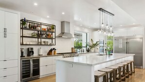 Een moderne keuken met witte kasten, een centraal eiland met barstoelen, roestvrijstalen apparatuur en diverse flessen en glazen op planken.