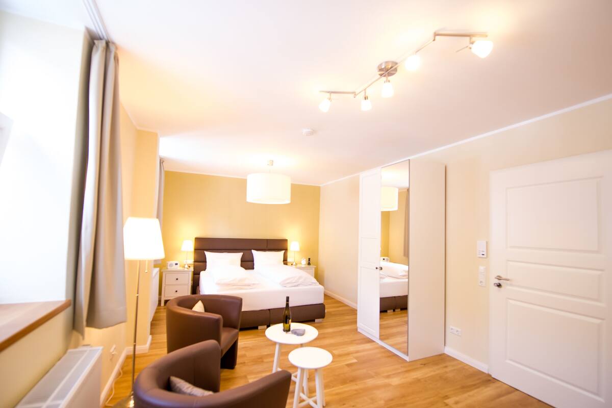 A hotel room with a bed, a sitting area with a chair and a small table, a nightstand with a lamp, and a door leading to another room.