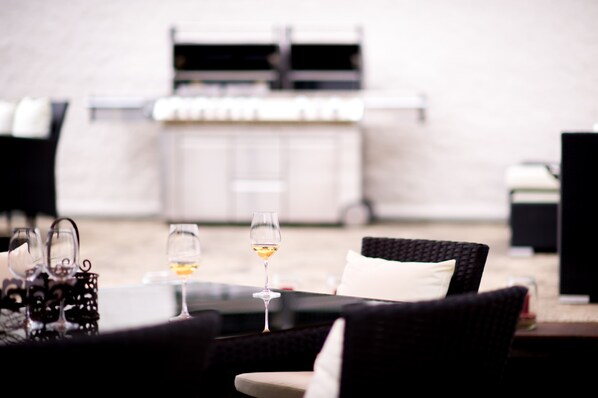 Uma área externa com assentos, cadeiras de vime e uma mesa de vidro, com dois copos de vinho.