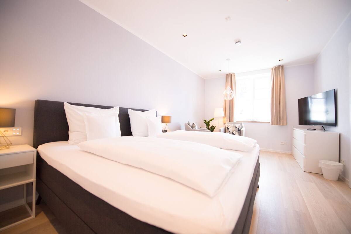 A modern hotel room with a large bed, a TV, a desk, and a window with curtains.